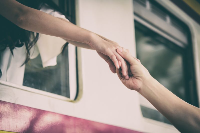 Two people holding hands as a train leaves. One is on the train and one is on the platform.