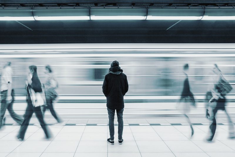 Person standing on a platform as a train and people pass by, giving a sense of isolation amongst busy places.