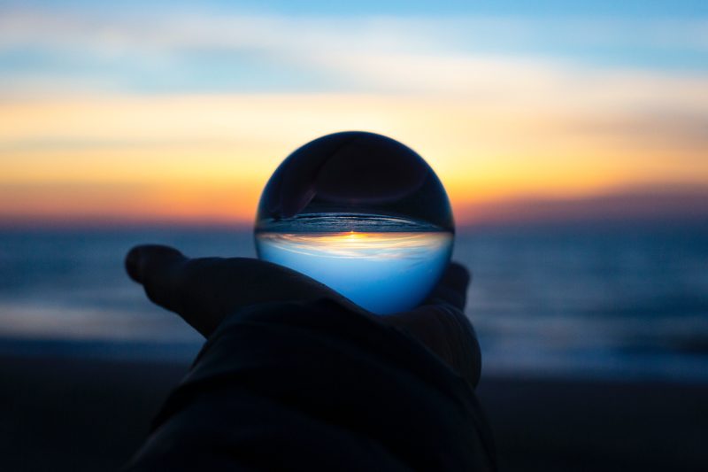 A hand holding a glass orb with a sunset over a beach inverted in it.